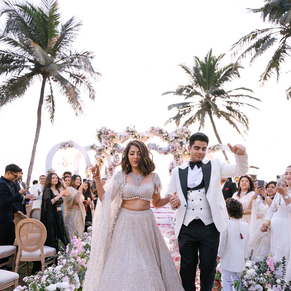 A bride in a pearl white lehenga and groom in a white tuxedo walking back down a petal strewn beach aisle framed by palms and a floral arch