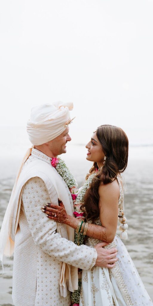A groom in a white turban and ivory sherwani holding his bride close on a misty beach at dusk, both in ivory wedding wear