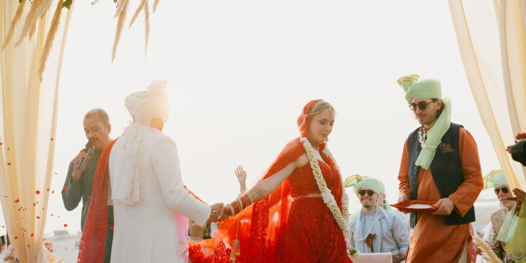 Guests in green turbans showering petals as the bride in a red and orange saree walks in holding the groom's hand at a Goa beach wedding
