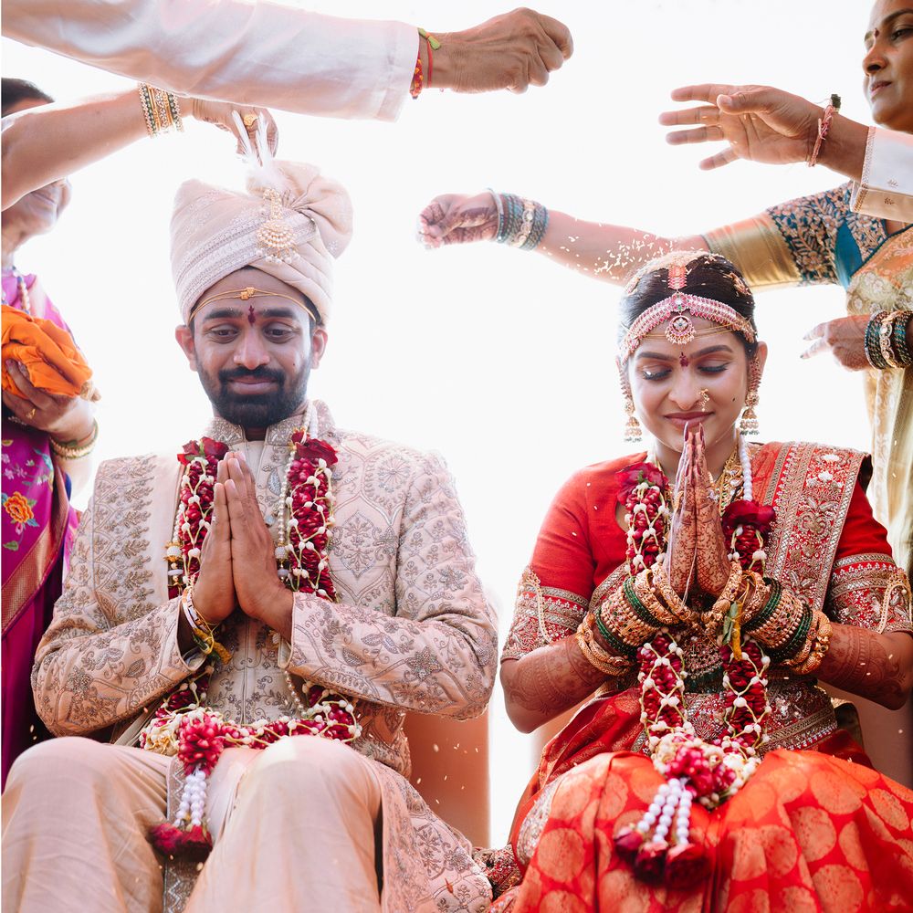 A groom in a cream sherwani and bride in a red Kanjeevaram saree sitting with folded hands as family showers rice over them
