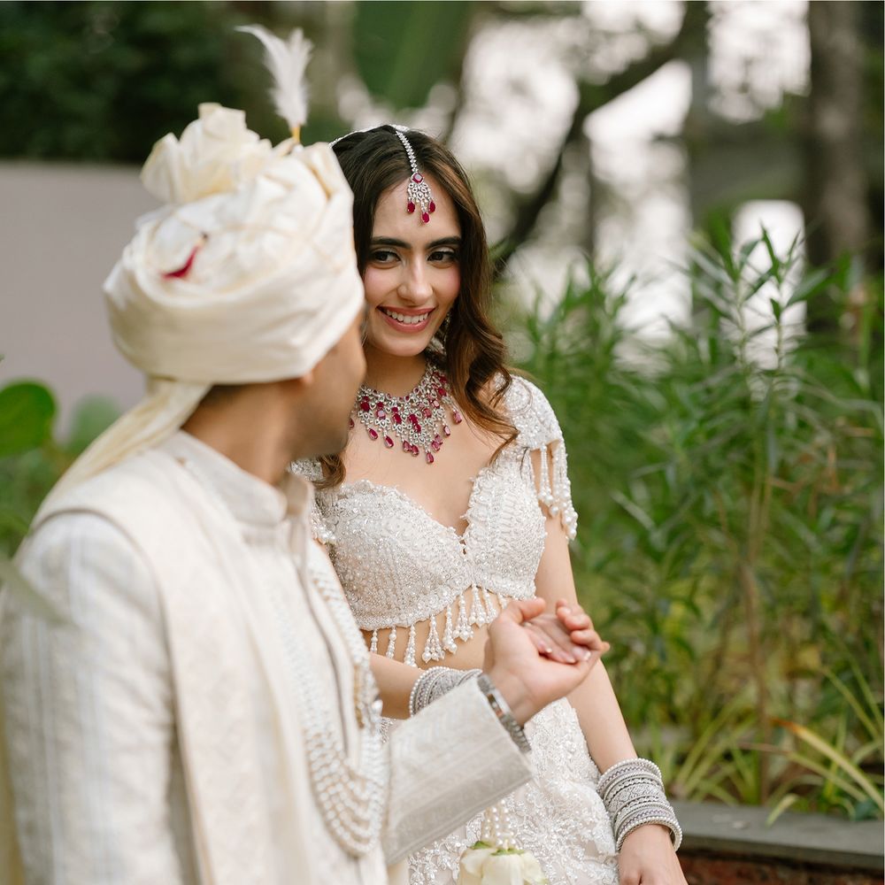 A groom in a white feathered turban and ivory sherwani holding hands with his bride in an ivory lehenga and ruby choker