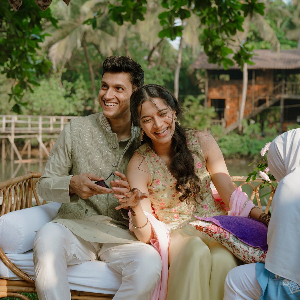 A couple laughing together on a cane seat surrounded by palms, he in a sage green kurta and she in a floral ivory lehenga