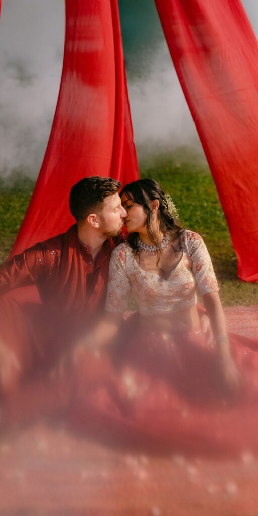A couple sitting close together under flowing red drapes on a lawn, the groom in a deep red kurta and the bride in a pastel embroidered lehenga