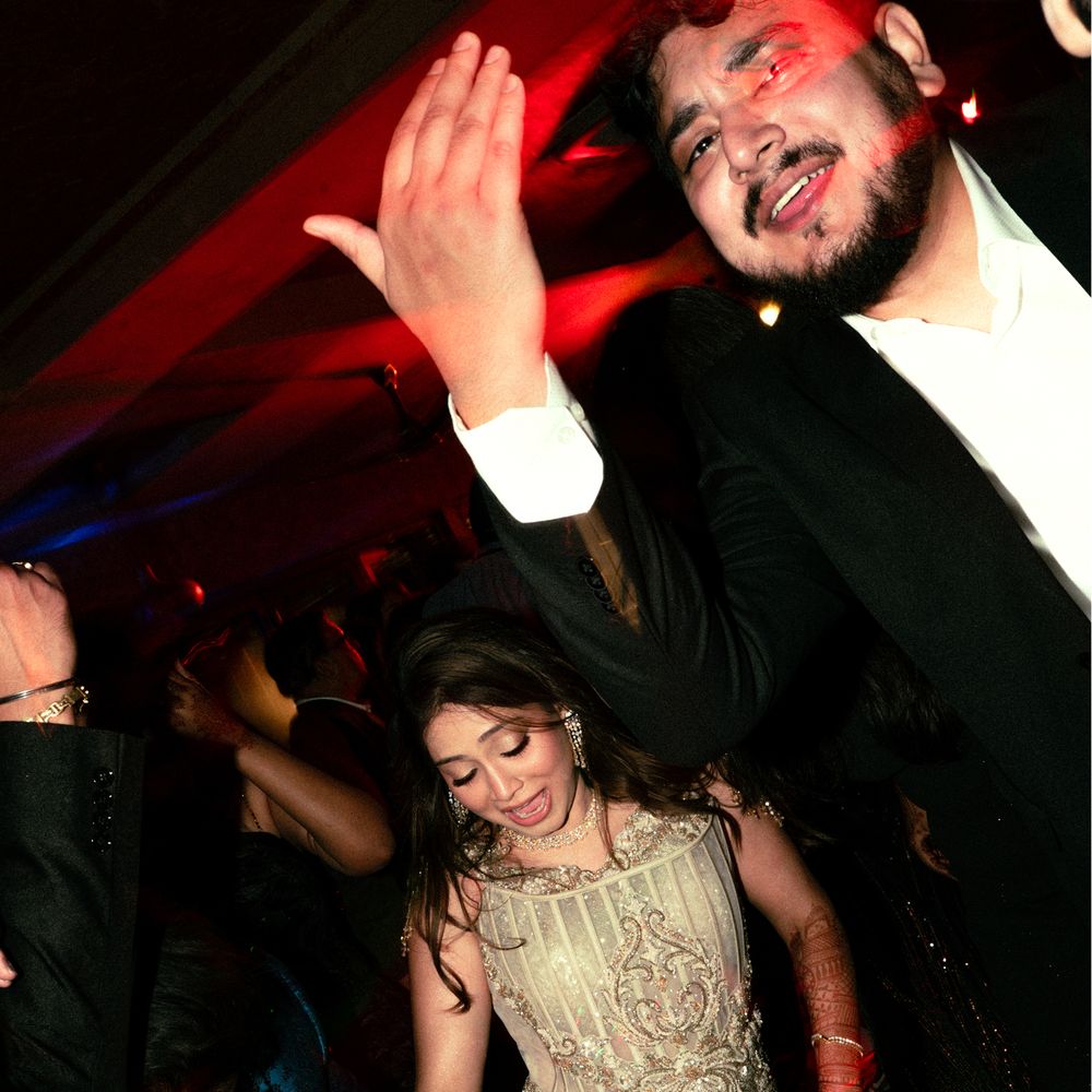 Guests dancing on a dark sangeet floor under red light, the bride in a gold sequin gown mid step