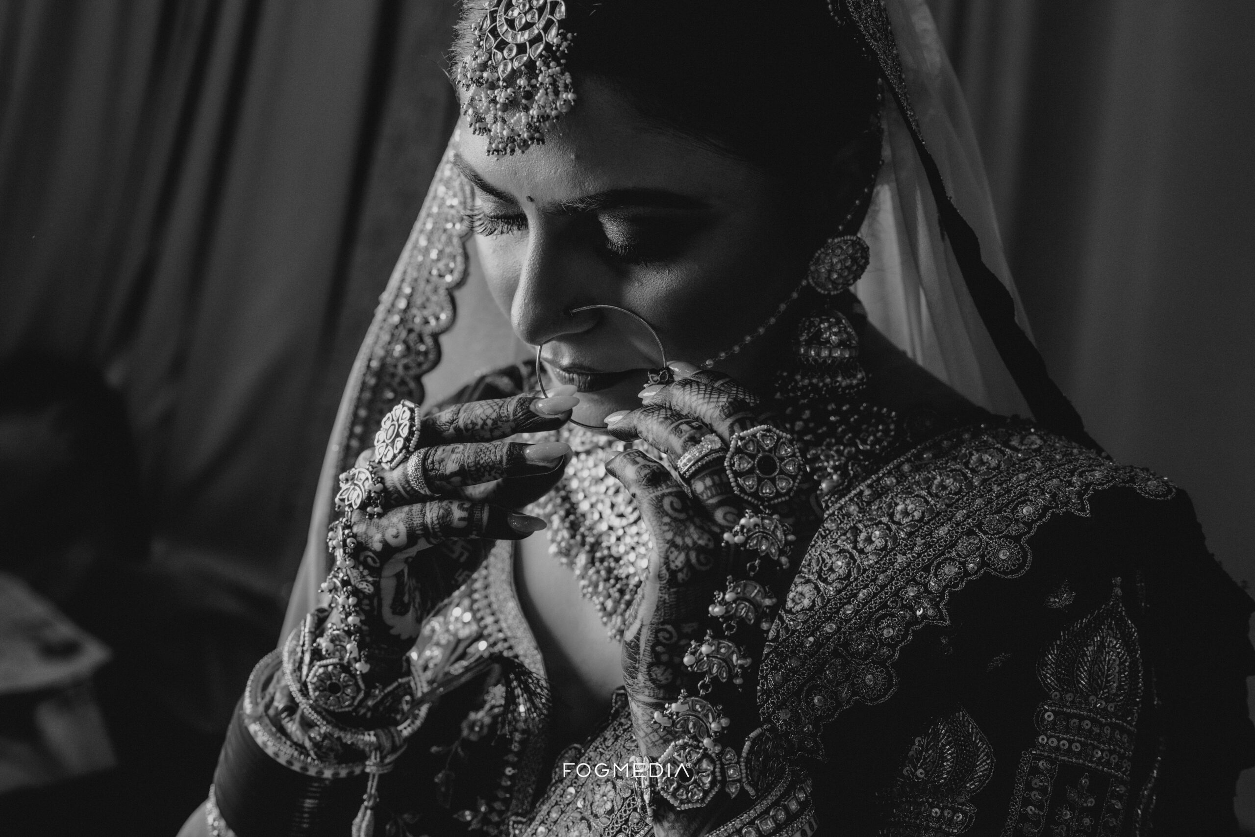 Black-and-white portrait of a bride looking down, wearing an embroidered veil and lehenga with a maang tikka, jhumka earrings, a large nose ring and mehndi across her hands