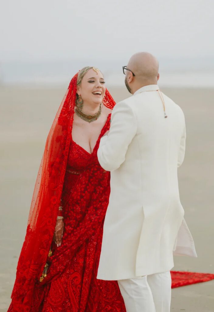 A bride in a red embroidered lehenga and veil laughing with her groom in a cream sherwani on a misty beach