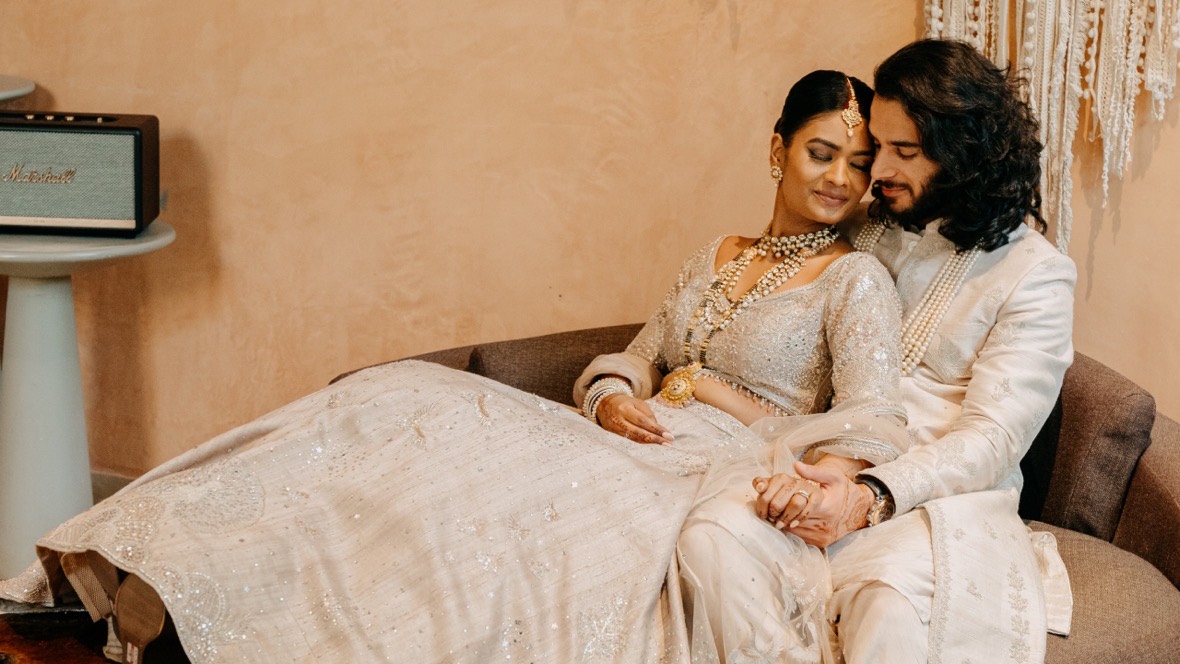 Ishitha and Shubh resting head to head on a curved couch against a warm peach wall, she in an ivory embroidered lehenga layered with pearl and gold necklaces, he in a cream sherwani, their hands clasped in her lap