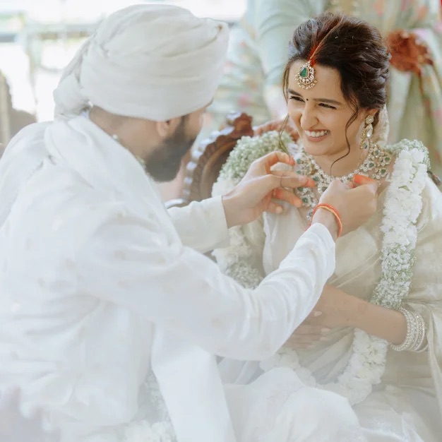 A groom in a white turban fastens the bride's necklace as she smiles, wearing a jasmine garland and a gold-and-green maang tikka