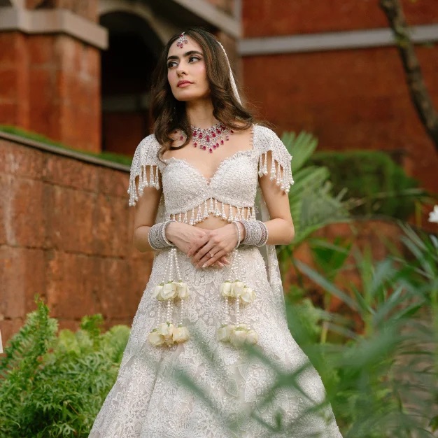 A bride in an ivory pearl-work lehenga stands before a red laterite heritage wall framed by tropical greenery
