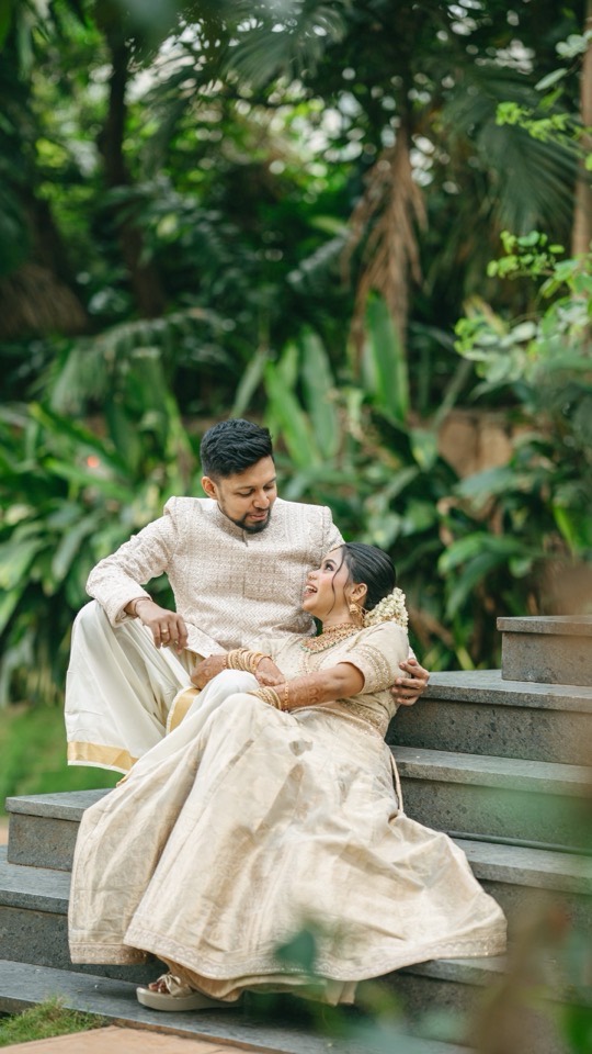 Goutam and Vaishnavi laughing together on stone steps banked by tropical greenery, he in a cream sherwani and mundu, she reclining against him in a gold-bordered cream sari with jasmine in her hair