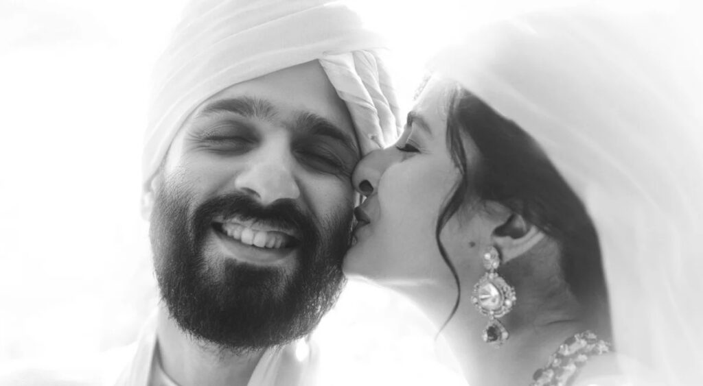 Wedding photography in Hyderabad — black-and-white close-up of a groom in a white turban smiling as his bride kisses his cheek, wearing an ornate chandelier earring